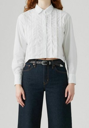 Vrouw draagt een witte blouse met knopen en kanten details, gestopt in donkerblauwe hoog getailleerde jeans met een zwarte riem.