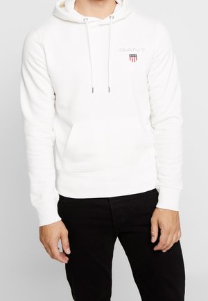 Sweat à capuche blanc en matériau doux avec une poche frontale, un cordon de serrage pour la capuche et un petit logo brodé sur la poitrine. Pantalons noirs contrastants.