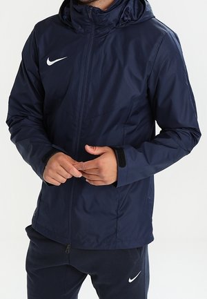 Mann trägt eine dunkelblaue Nike Kapuzenjacke und passende Hose mit weißen Nike Swoosh-Logos, Hände vor dem Körper verschränkt.