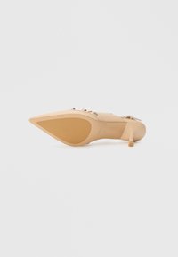 Zapato beige de tacón alto con punta estrecha, material liso, detalles discretos de metal y tacón de aguja delgado. Suela de color marrón claro con textura mínima.