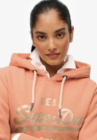 Sweat à capuche pêche avec cordons, arborant un logo "SuperDry" doré. Porté sur une chemise blanche à col, avec une texture lisse et une coupe décontractée.