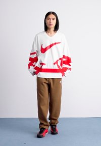 Person iført hvid langærmet skjorte med rød abstrakt Nike-logo, brune bukser og sort-røde sneakers, der står op imod en helt hvid væg.
