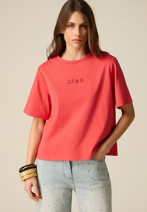 Junge Frau trägt ein korallenrotes Kurzarmshirt mit dem Schriftzug "ciao" und hellblaue Jeans mit kleinen Strasssteinen, accessoriert mit klobigen Armbändern.