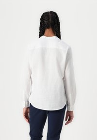 Camisa de lino blanca con un ajuste relajado, mangas largas y puños con botones. Presenta un detalle de yugo en la parte superior de la espalda y un dobladillo curvado.