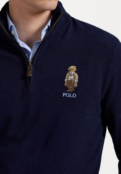 Navy zip-up sweater with a knitted texture, featuring an embroidered bear in a tan suit and "POLO" in light blue underneath.