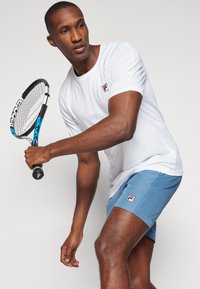 Valge sportline särk Fila logo ja helesinised lühikesed püksid. Teema hoiab tennisereketit, näidates aktiivset ja dünaamilist poosi.
