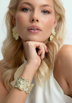 Örhängen i form av guldkvistar och ett texturerat guldarmband bärs av en modell. Smyckena har intrikata detaljer och en reflekterande yta.