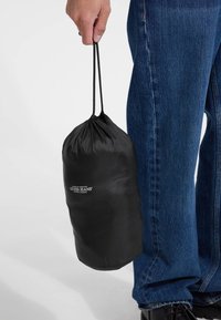 Crna torba na vezanje izrađena od laganog materijala, cilindričnog oblika, sa bijelim logotipom "Guess Jeans". Drži je ruka naslonjena na plave traperice.