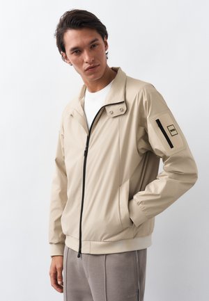 Άνδρας που φοράει μπεζ ζακέτα με φερμουάρ και τσέπη με πατσαλίκι στο μανίκι, καθώς και μπεζ παντελόνι, στέκεται μπροστά σε απλό φωτεινό φόντο.