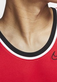 Red athletic tank top with black and white ribbed neckline. Features Nike logo in black on the left side, mesh texture for breathability.