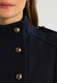 Abrigo de lana azul marino con un alto cuello, botones dorados detallados y costuras estructuradas para un aspecto entallado.