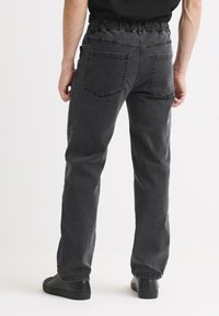 Mies, jolla on mustat joustovyötäröiset farkut ja mustat lenkkarit, seisoo selin yksinkertaista valkoista taustaa vasten.