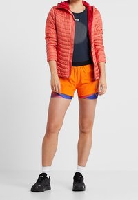 Röd quiltad jacka över en marinblå t-shirt med en rund grafisk design, kombinerad med orange shorts med blå accenter och svarta sportskor.