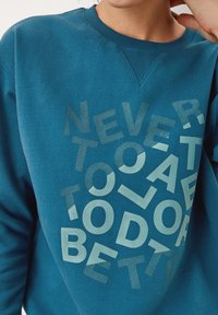 Felpa teal con collo rotondo; lettering bianco testurizzato in più font sul davanti che recita "NON È MAI TARDI PER FARE MEGLIO." Tessuto morbido.