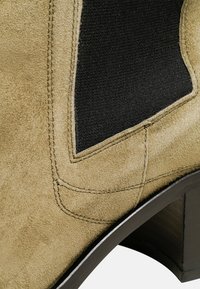 Beige suède enkellaars met een zwart elastisch paneel, een taps toelopende houten hak en zichtbare stiksels langs de naden.