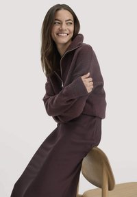 Bordeaux, rits-vet met relatief lange mouwen en een hoge kraag, gecombineerd met een bijpassende, strakke, lange rok. Zachte textuur en relaxte pasvorm.