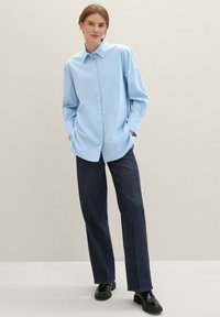 Femme debout dans une chemise oversize bleu clair, un jean large noir, des chaussures noires, les mains dans les poches, devant un fond beige uni.