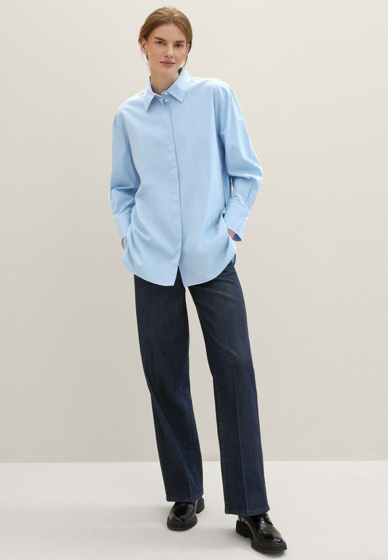 Kobieta stojąca w jasnoniebieskiej, oversize'owej koszuli, ciemnych, szerokich dżinsach i czarnych butach, z rękami w kieszeniach na jednolitym beżowym tle.