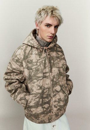 Jonge persoon met kort blond haar, draagt een beige en bruin capuchonjack met een abstract patroon en heeft de handen in de zakken.