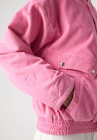 Rosa kordfløyelsjakke med ribbet tekstur, elastisk midje, trykknapp-lomme og tettsittende ermer. Har en uformell, boksformet fasong.