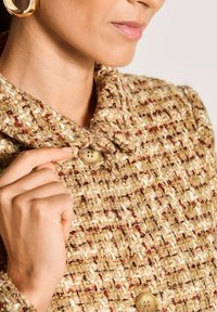 Vrouw die de kraag van een textuurrijke beige en bruine geweven jas met grote knopen aanpast, met een gouden ringoorbel.