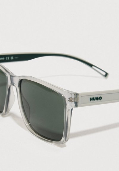 Lunettes de soleil carrées transparentes avec des verres vert foncé, dotées d'une branche blanche marquée "HUGO" en lettres vertes.