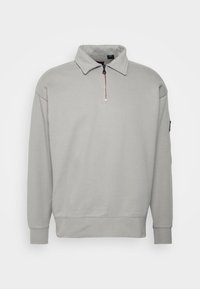 Sudadera de color gris claro con cuello con cremallera, hecha de un tejido suave. Presenta mangas largas y un ajuste relajado, con una textura sutil.