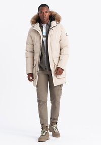 Béžová zateplená parka s kapucí podšitou kožešinou, přední kapsy, v kombinaci se šedou košilí, béžovými cargo kalhotami a hnědými botami se světlými tkaničkami.