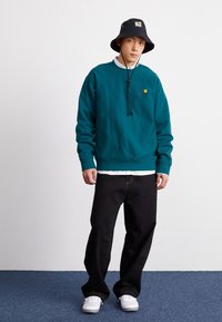 Teal sweatshirt med rund hals och färgglad logotyp, kombinerad med svarta vida byxor och en svart bucket hat. Vita sneakers fulländar looken.