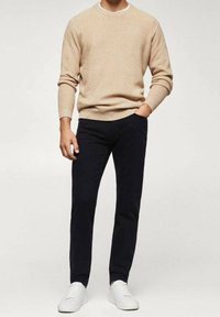 Pull en tricot beige avec un col rond, associé à un pantalon bleu marine. Des baskets blanches complètent le look. Texture lisse et design ajusté.