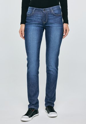 Nő karcsúsított kék farmert, fekete hosszú ujjú felsőt és fekete alacsony szárú tornacipőt visel, világos, egyszínű háttér előtt áll.