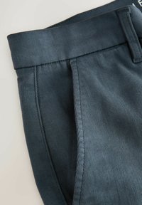 Sötétszürke szövet nadrágzseb látható varrásokkal és széles derékpánttal, világos háttér előtt.