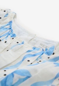 Biela tkanina s modrými vlnkovými vzormi a čiernymi bodkami, so zhrnutým materiálom na výstrihu. Na štítku je nápis "NEXT, MADE IN."