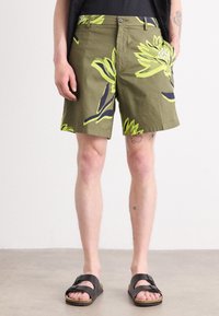 Homme portant un short vert olive avec de grandes impressions florales jaunes, des sandales noires et une chemise noire, debout sur un fond gris clair.