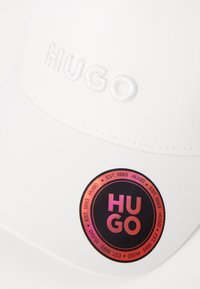Hvid kasket med broderet "HUGO" på forsiden og et rundt, multikolor label med "HUGO" og details om etableringen.
