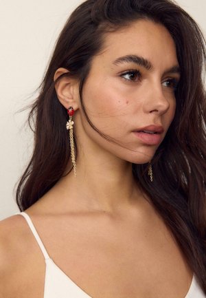 Młoda kobieta o długich ciemnych włosach, ubrana w biały top i wiszące kolczyki z motywem biedronki oraz złotym łańcuszkiem, patrząca na bok.