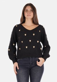 Maglione nero con scollo a V, con motivi a cuore dorati, polsini e orlo a coste. Il tessuto appare morbido eTexture. Indossato con jeans scuri.