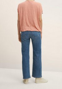 Personne portant un haut rose texturé à manches courtes, un jean bleu droit et des baskets blanc cassé, debout devant un fond uni.