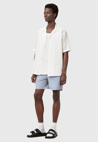 Camicia bianca a maniche corte con texture verticale, pantaloni corti grigi leggeri, sandali neri e calzini bianchi. Design semplice con chiusura a bottone.