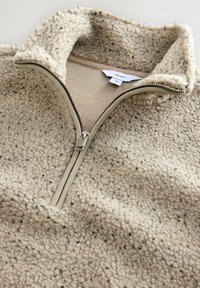 Beige fleece sweatshirt met een hoge kraag en een rits aan de voorkant. Textuurstof met een zachte werking, voorzien van kleine donkere vlekken en lichtbruin accent.