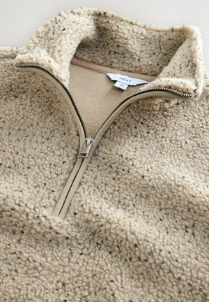 Sudadera de poliéster beige con cuello alto y cierre de cremallera. Tejido texturizado con una sensación suave, que presenta pequeñas manchas oscuras y acentos de un tono marrón claro.