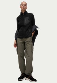 Chaqueta de forro polar negra con cuello alto y bolsillo frontal, combinada con pantalones cargo oliva que tienen bolsillos laterales, y zapatos deportivos negros.