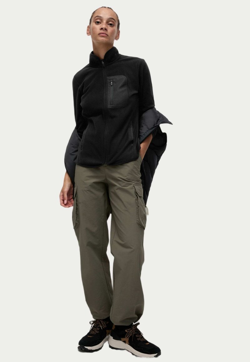 Chaqueta de forro polar negra con cuello alto y bolsillo frontal, combinada con pantalones cargo oliva que tienen bolsillos laterales, y zapatos deportivos negros.