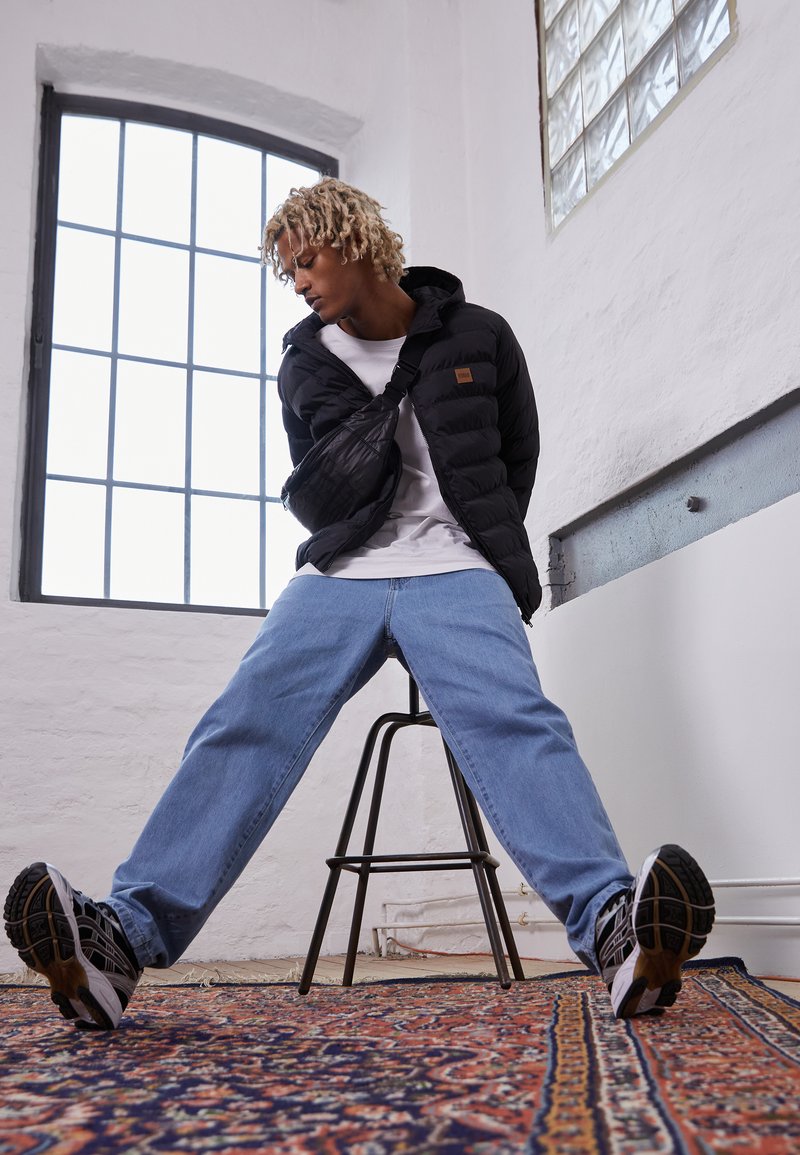 Sort pufferjakke med hætte, hvid t-shirt, lyseblå jeans og sorte sneakers, poseret foran et stort vindue og et mønstret tæppe.