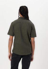 Camisa de manga corta verde oliva con cuello, que presenta una textura suave y un dobladillo curvado, combinada con pantalones negros.