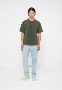 Olijfgroen T-shirt met korte mouwen en een licht oversized pasvorm, met een kleurrijke grafische print op de voorkant. Gecombineerd met lichtblauwe jeans en witte sneakers.