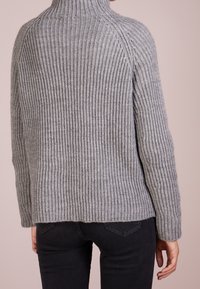 Eine Person trägt einen grauen, gerippten Strickpullover mit hohem Kragen und schwarze Hosen. Sie wird von hinten vor einem schlichten Hintergrund gezeigt.