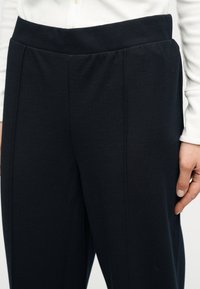 Κοντινό πλάνο ατόμου που φοράει μαύρο παντελόνι με ψηλή μέση και λευκό μακρυμάνικο τοπ, με το χέρι χαλαρό στο πλάι.