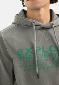 Gray hoodie with drawstring, featuring green printed text. Made of soft fabric, includes a front pocket and decorative stitching details.