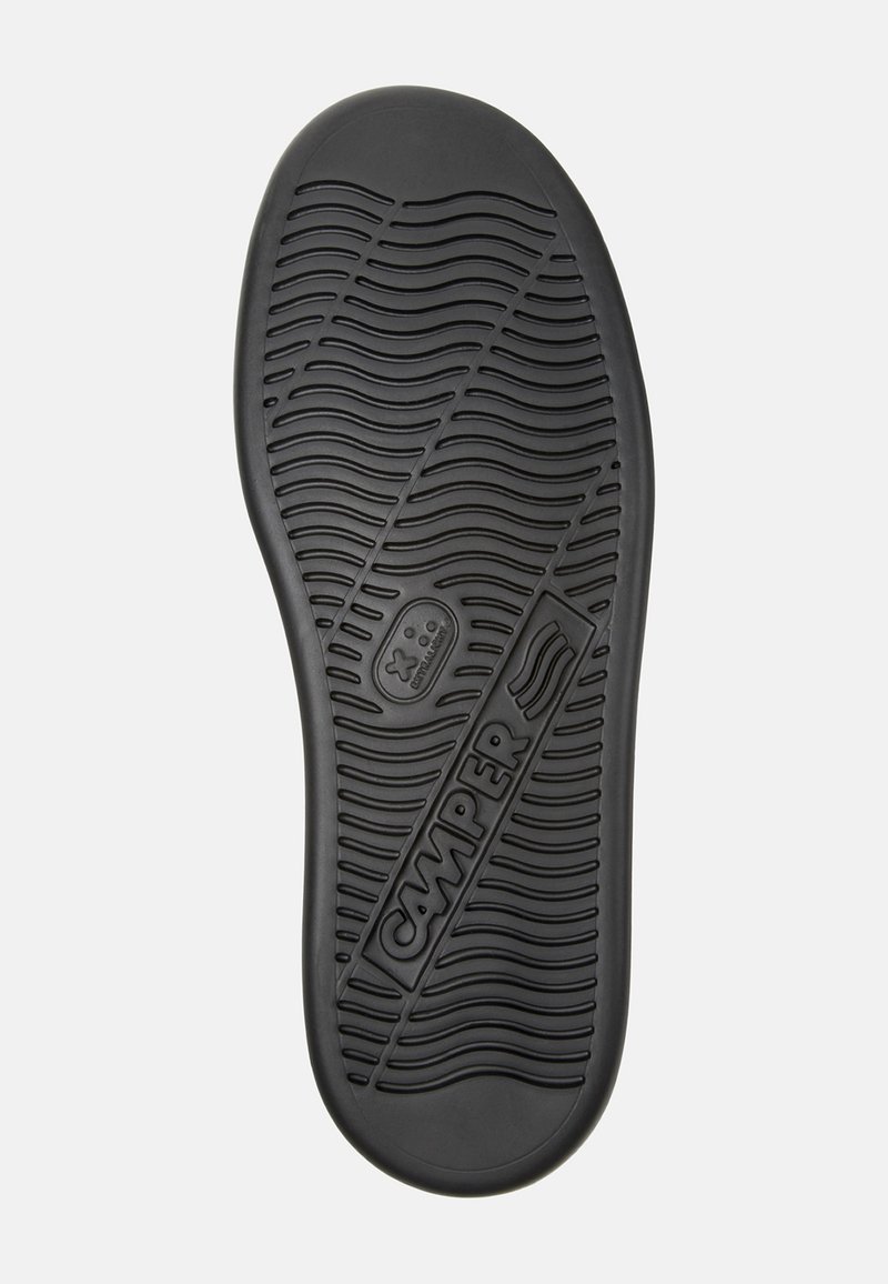 Black rubber shoe sole with textured wave pattern, featuring a circular logo and the brand "CAMPER" embossed. Smooth finish, slightly curved edges.